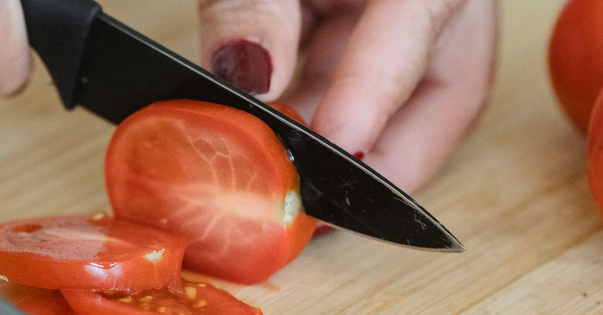 Un gros plan de mains tranchant des tomates fraîches et mûres sur une planche à découper en bois avec un couteau bien aiguisé.