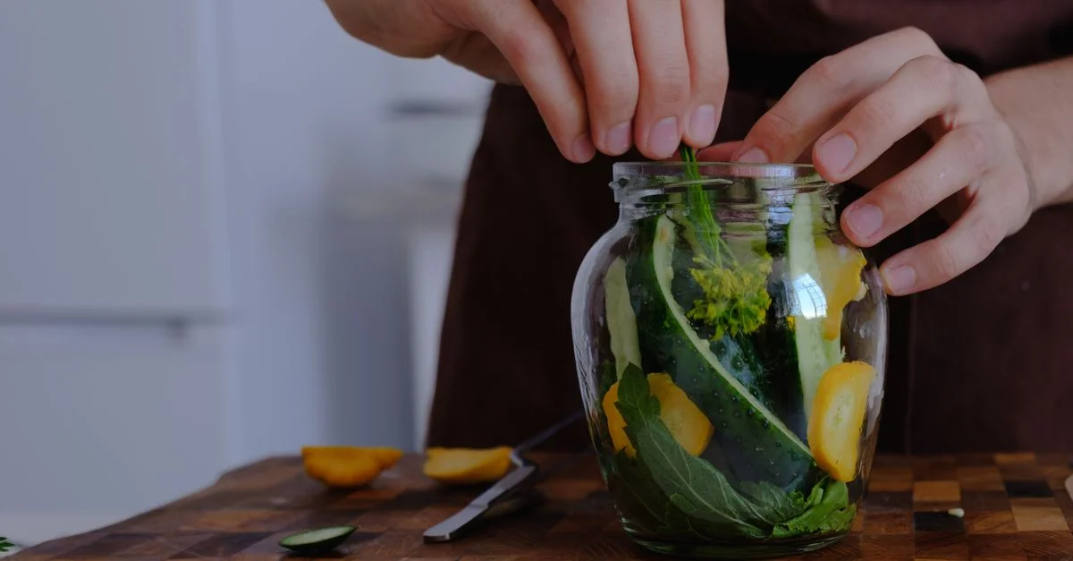 Des mains organisant des légumes frais pour la mise en conserve dans un pot en verre sur un plan de travail en bois.