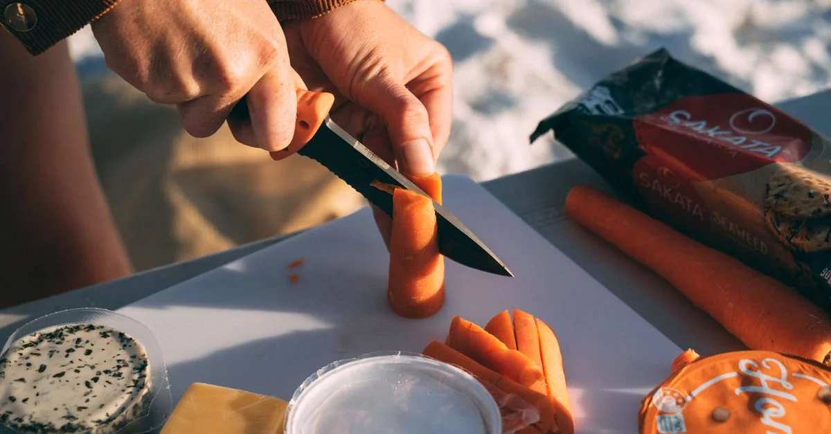 Des mains tranchant des carottes sur une planche à découper avec du fromage et des collations, parfait pour les pique-niques en plein air.