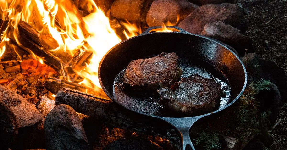 Des steaks grésillant dans une poêle en fonte au-dessus d'un feu de camp entouré de bûches et de pierres.