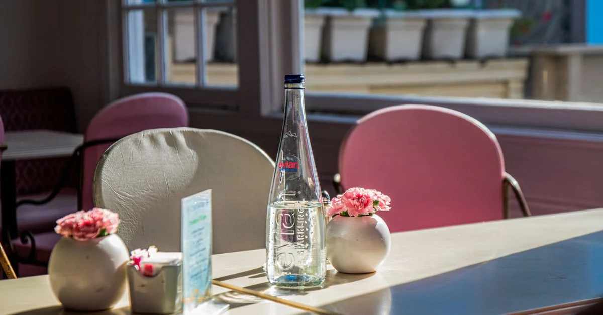 La lumière du soleil projette des ombres sur une table de café avec une bouteille d'eau, des fleurs roses et des chaises.