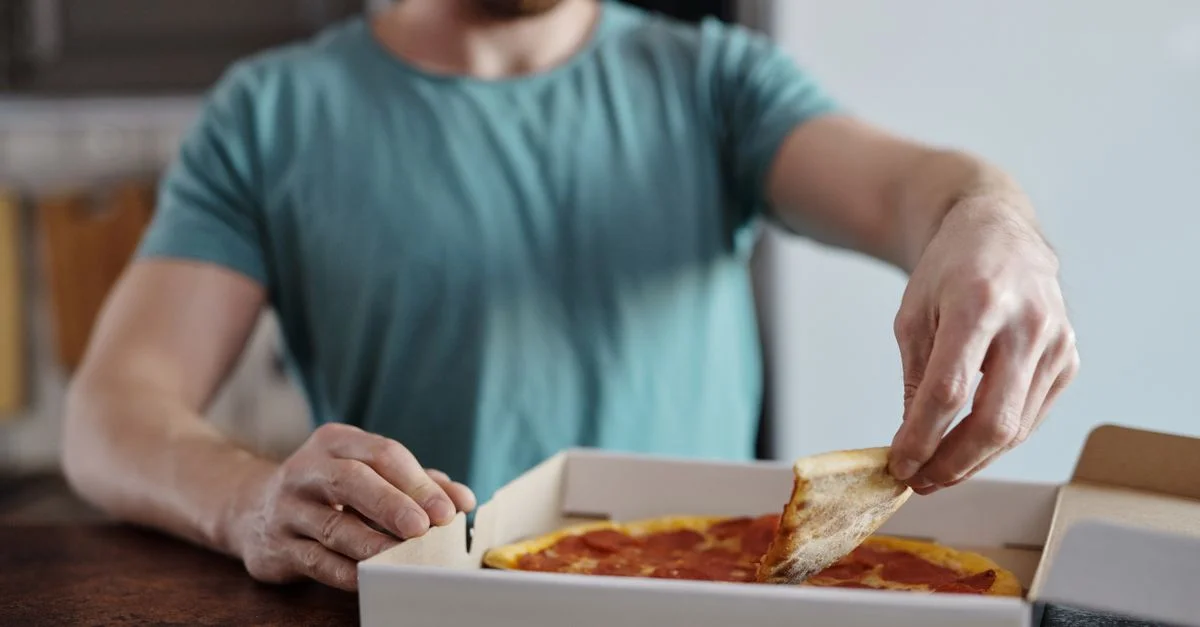 Une personne en chemise turquoise prenant une part de pizza pepperoni d'une boîte à l'intérieur.