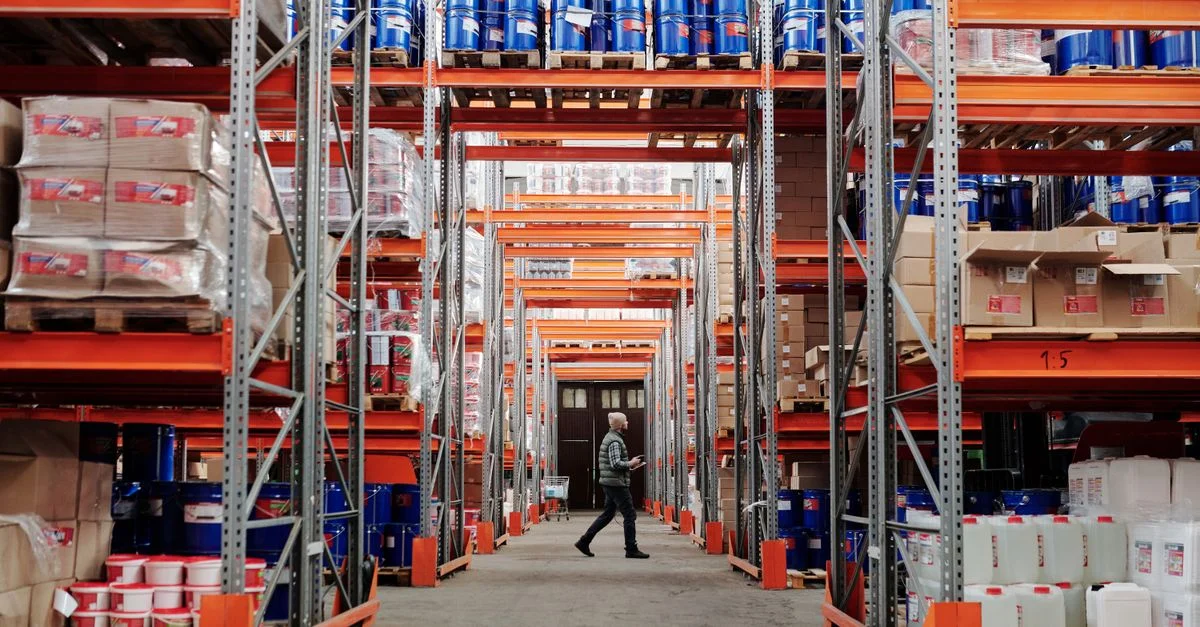 Un homme marchant à travers un grand entrepôt industriel avec des étagères empilées de biens et de produits.
