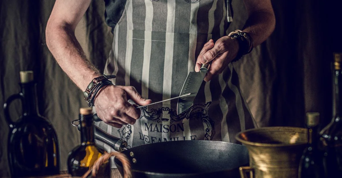 Un chef aiguise un couteau dans un cadre de cuisine rustique, portant un tablier rayé.