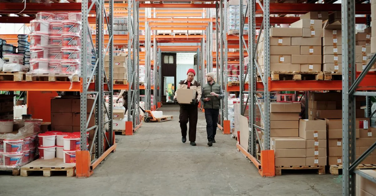 Deux travailleurs manipulent un colis dans un entrepôt spacieux entouré d'étagères chargées de boîtes et de produits.