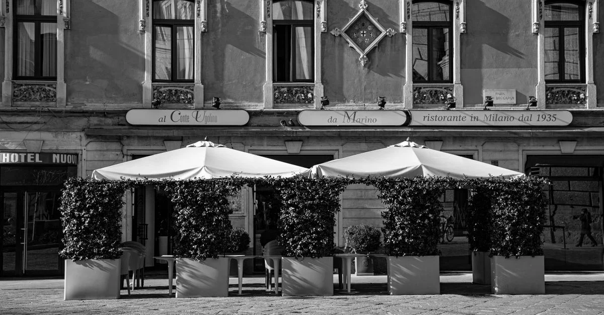 Photo en noir et blanc d'un café italien historique avec des sièges extérieurs et des parasols par une journée ensoleillée.