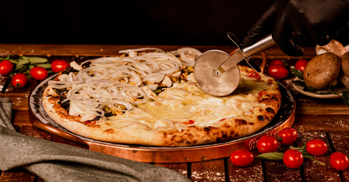 Pizza artisanale avec des oignons tranchés par une main gantée, entourée de tomates cerises fraîches.