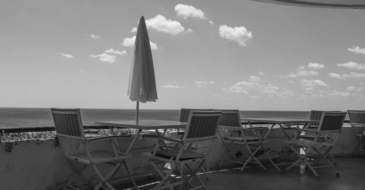 Vue en noir et blanc d'une terrasse en bord de mer avec des chaises vides et un parasol.